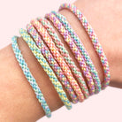 A close-up of a wrist wearing six braided elastic bracelets in pastel rainbow colors, including pink, blue, yellow, green, and purple, against a soft light pink background.