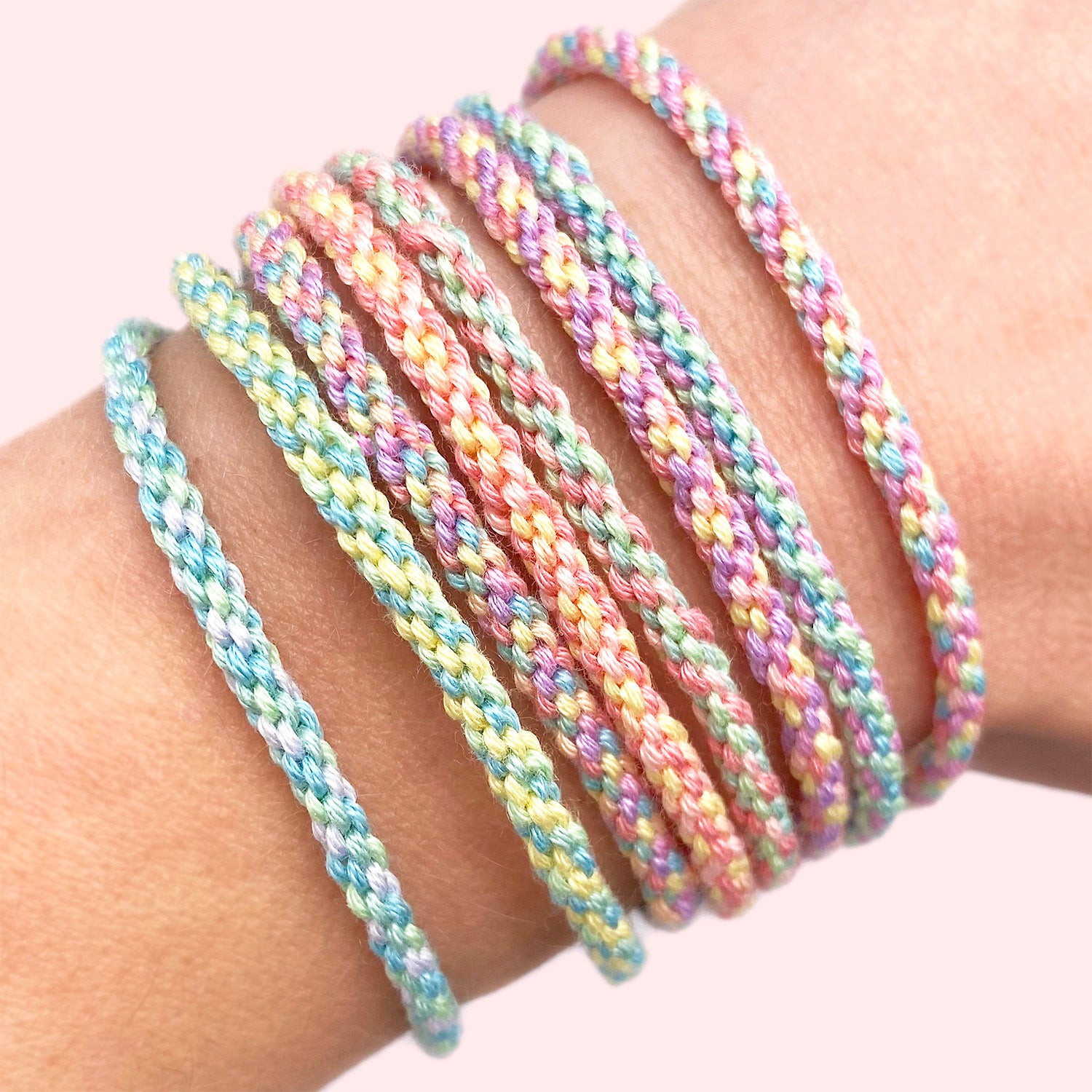 A close-up of a wrist wearing six braided elastic bracelets in pastel rainbow colors, including pink, blue, yellow, green, and purple, against a soft light pink background.