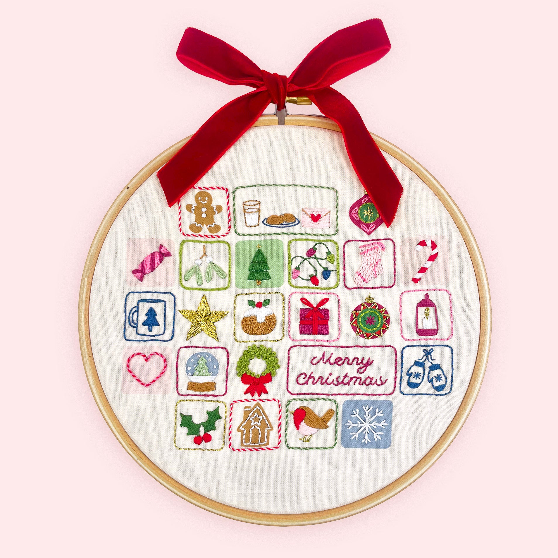 Embroidery advent calendar. A circular embroidery hoop with a wooden frame holds a white fabric panel featuring colorful Christmas-themed stitched icons and a red 'Merry Christmas' script. A red velvet ribbon is tied in a bow at the top for hanging.
