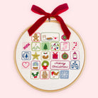 Embroidery advent calendar. A circular embroidery hoop with a wooden frame holds a white fabric panel featuring colorful Christmas-themed stitched icons and a red 'Merry Christmas' script. A red velvet ribbon is tied in a bow at the top for hanging.