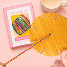 A craft kit box labeled 'Lazy May Sewing Club' sits on a pink background next to a large circular wooden disc with cutout sections and a braided bracelet threaded through the center. A hand is holding the bracelet, and a cup is partially visible at the top right.