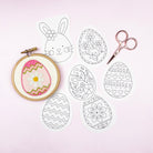 Embroidery hoop with a completed Easter egg design, scissors, and coloring pages on a light pink background