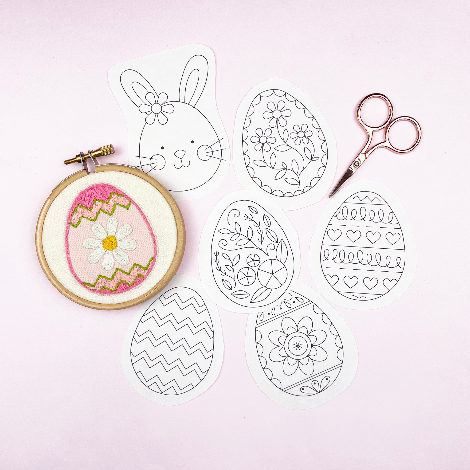 Embroidery hoop with a completed Easter egg design, scissors, and coloring pages on a light pink background