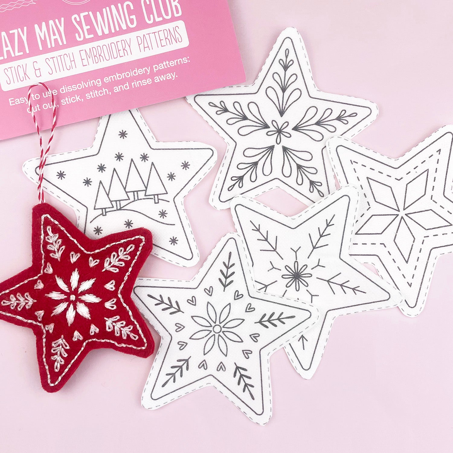 Six star-shaped embroidery pattern pieces with snowflake and tree designs are arranged on a light pink background, with a pink package card reading 'Lazy May Sewing Club Stick & Stitch Embroidery Patterns' and a red embroidered star ornament hanging from one piece.