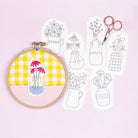Embroidery hoop with yellow fabric and floral design, next to white paper patterns of vases and scissors on a pink background.