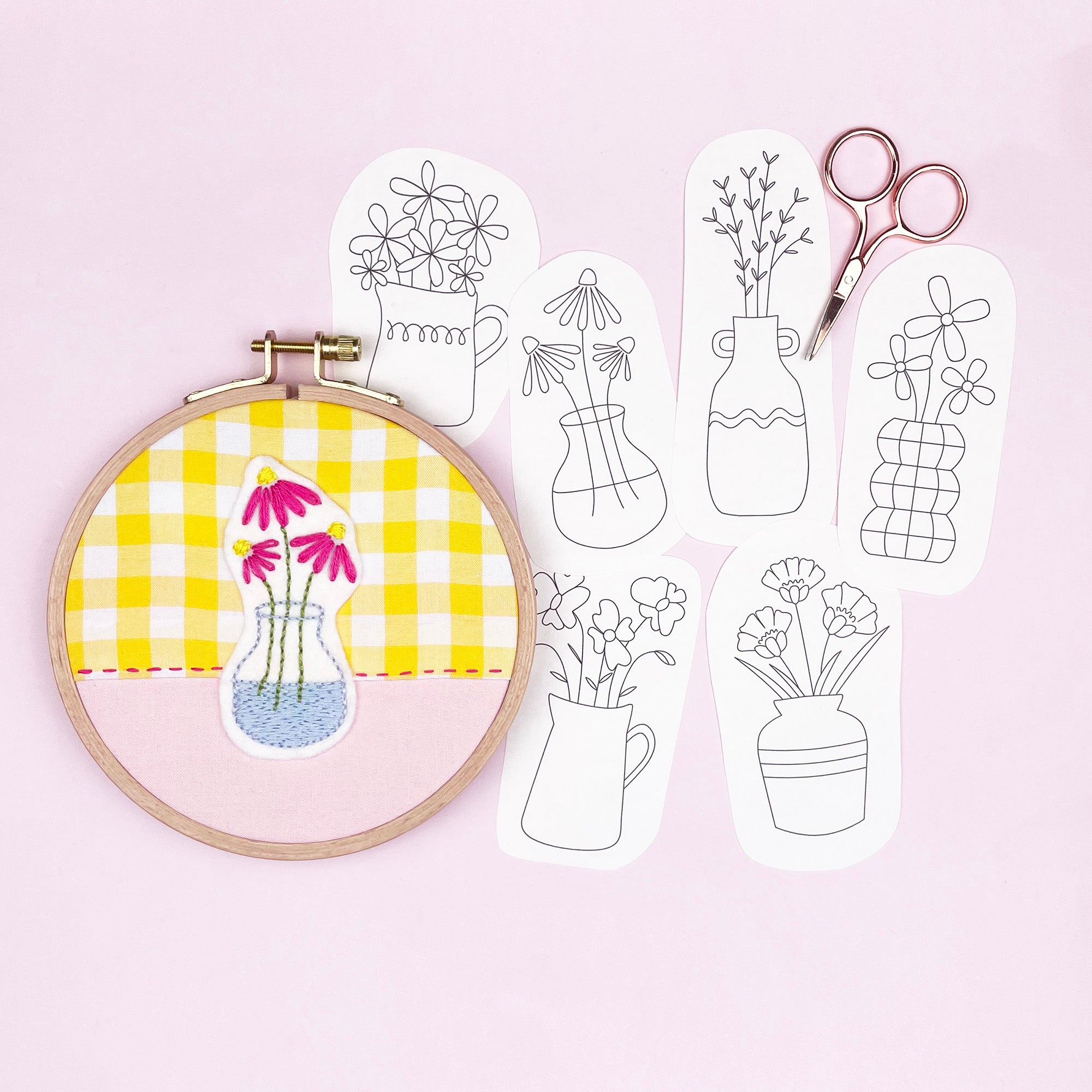 Embroidery hoop with yellow fabric and floral design, next to white paper patterns of vases and scissors on a pink background.