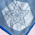 A close-up of a light blue woven textile with a white stitched star motif and blue fringe along the edge, shown against a pale pink background.
