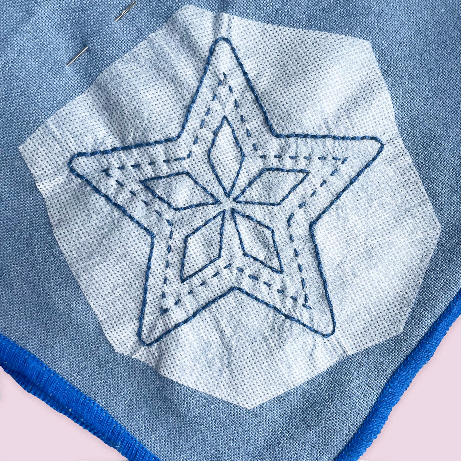 A close-up of a light blue woven textile with a white stitched star motif and blue fringe along the edge, shown against a pale pink background.