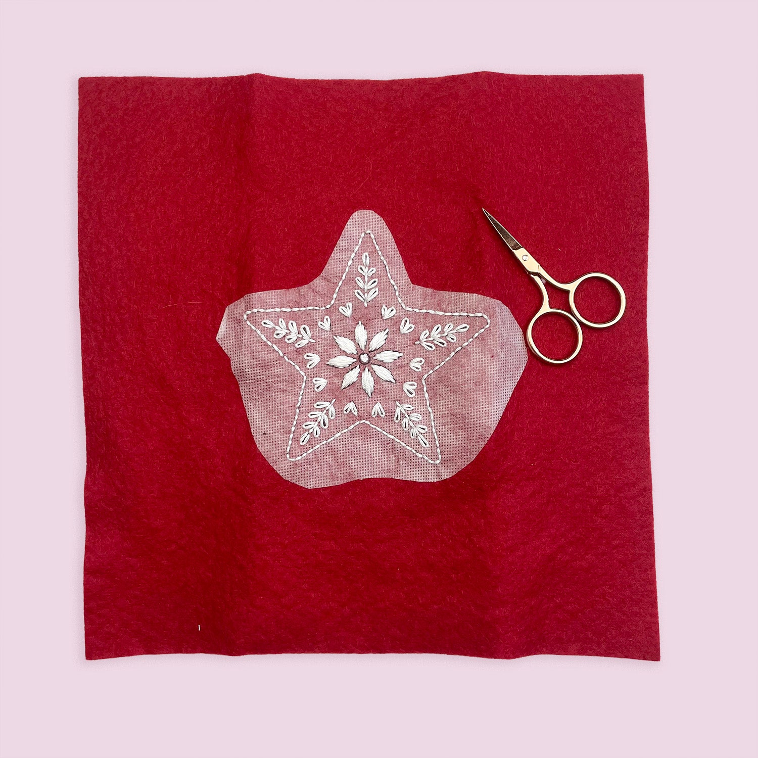 A square piece of deep red fabric with a star-shaped stick and stitch embroidery patch, styled with a pair of gold-handled scissors on a light pink background.