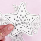 A hand holds a white star-shaped stick and stitch embroidery pattern with black stitched outlines and a simple tree landscape design, with other similar star ornaments blurred in the background on a light pink surface.