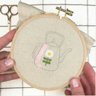 Embroidery hoop with a teapot and flower design held by a hand on a checkered background.