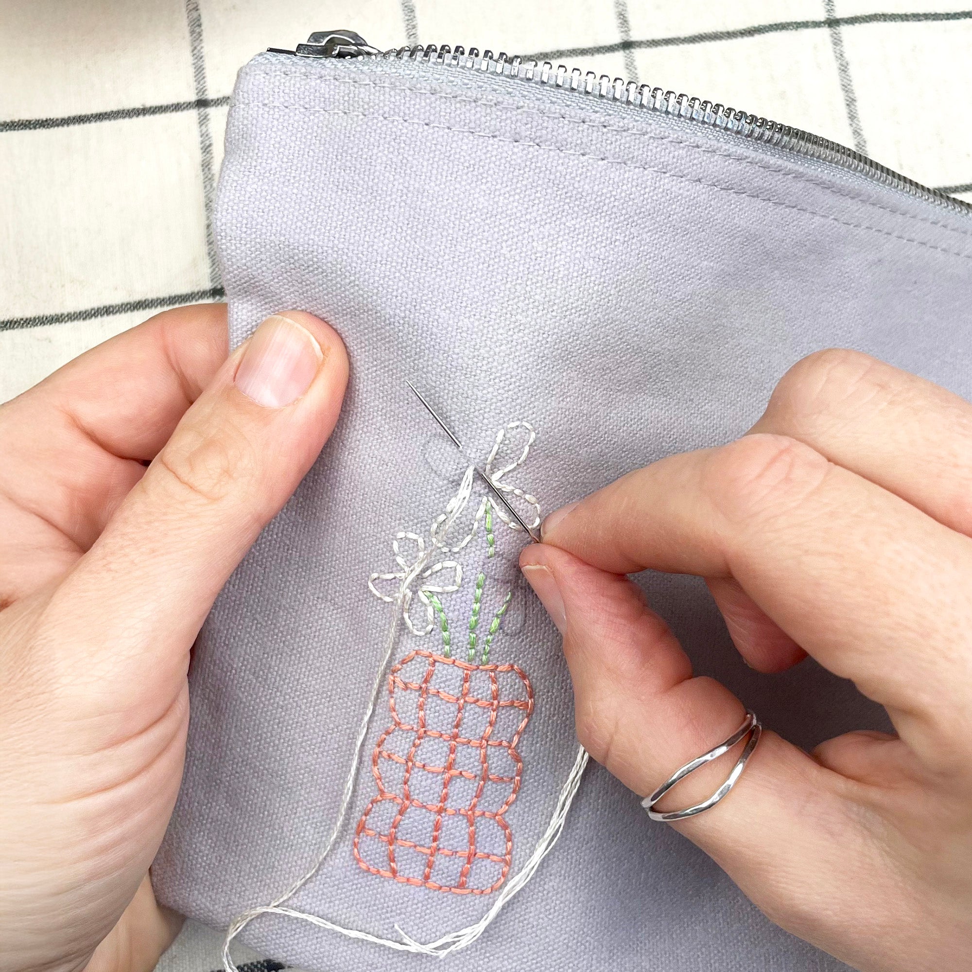 Person embroiderying a design on a gray fabric pouch with a needle and thread.