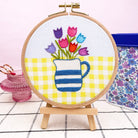 Embroidery hoop with colorful tulips and a striped pitcher on a yellow checkered background.