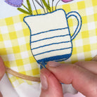 Embroidery of a blue and white striped mug with flowers on a yellow and white checkered fabric.