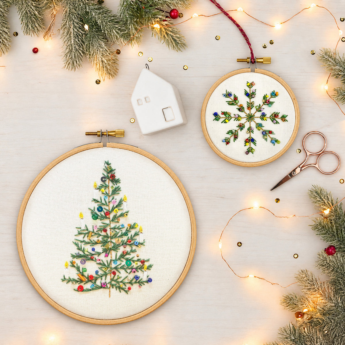 Two round embroidery hoops displayed on a light wood surface, surrounded by holiday greenery, string lights, scissors, and a small white house ornament. One hoop shows a decorated Christmas tree, and the other shows a colorful snowflake.