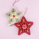 Two embroidered felt star Christmas ornaments hanging against a light pink background. One star is cream with multicolor floral embroidery, and the other is red with white floral embroidery.