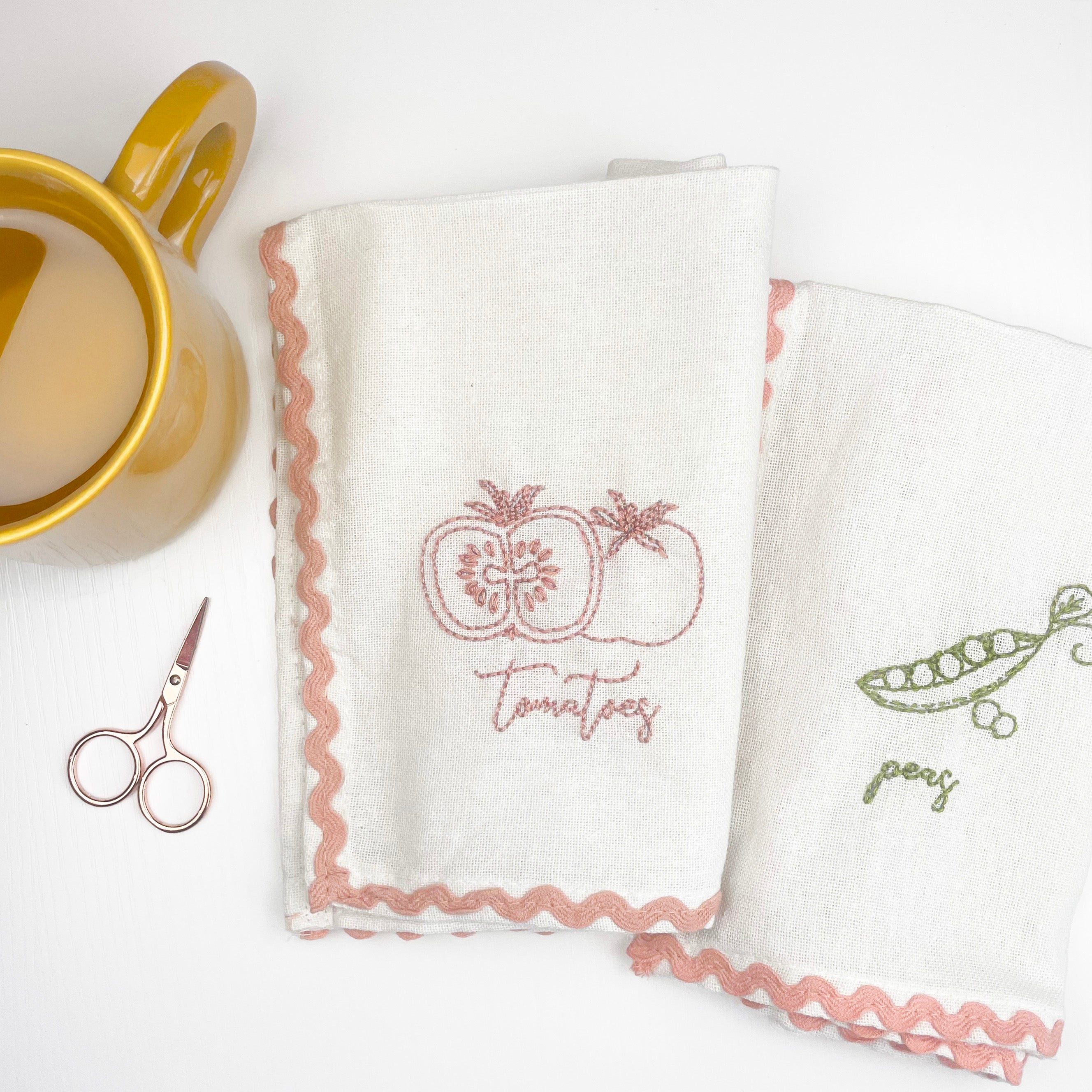 Two embroidered towels with vegetable designs and text next to a mug and scissors on a white background