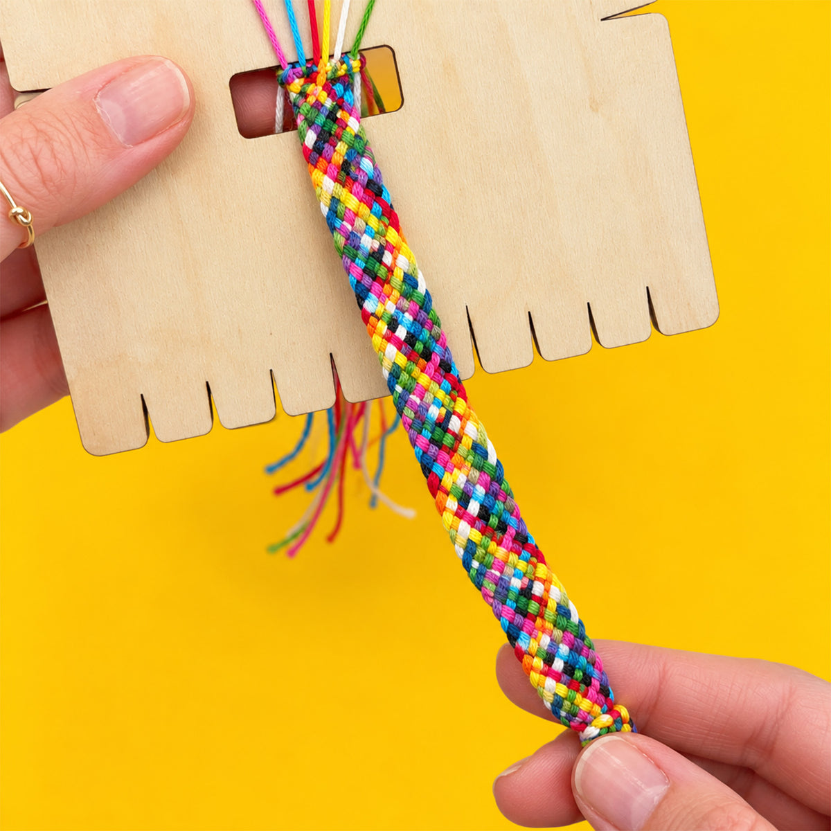 A close-up of a hand holding a colorful braided friendship bracelet in front of a wooden display stand, with the other hand holding the stand against a bright yellow background.