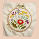A circular embroidery hoop holding a floral needlepoint or embroidered design on a cream fabric background, photographed against a soft peach-pink backdrop.